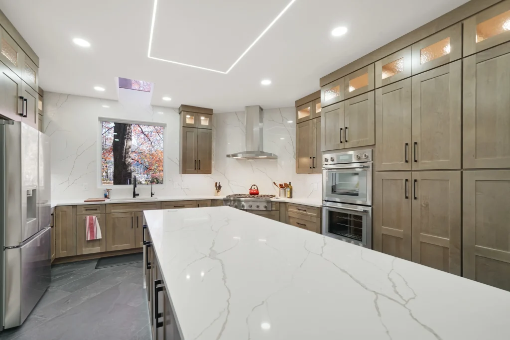 Modern kitchen with light wood cabinets, stainless steel appliances, marble island countertop, under-cabinet lighting, and large window above the sink.