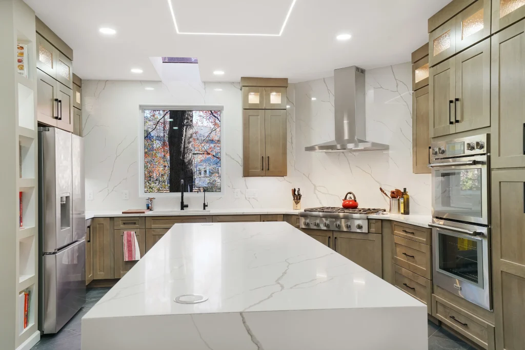 Modern kitchen with wood cabinetry, marble countertops, stainless steel appliances, a large island, and a window showing trees outside.
