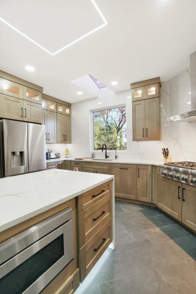 Modern kitchen with light wood cabinets, stainless steel appliances, marble countertops, and a large window overlooking trees. Recessed and under-cabinet lighting illuminate the space.