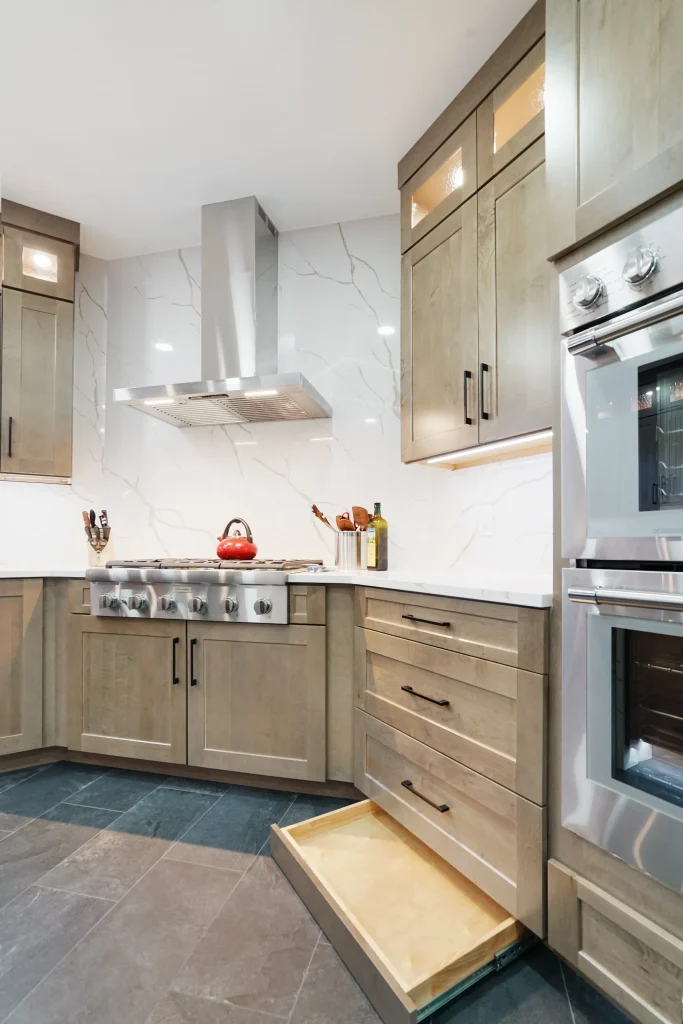 Modern kitchen with light wood cabinets, stainless steel appliances, a gas stove with a red kettle, and a pull-out drawer open at the bottom. Marble backsplash and dark tile flooring.