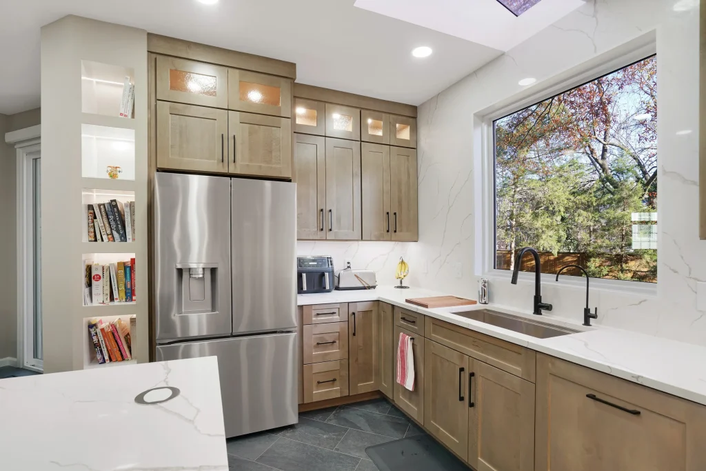 Modern kitchen with stainless steel refrigerator, built-in oven, light wood cabinets, large window, and a marble countertop. Bookshelves are built into the cabinetry near the refrigerator.