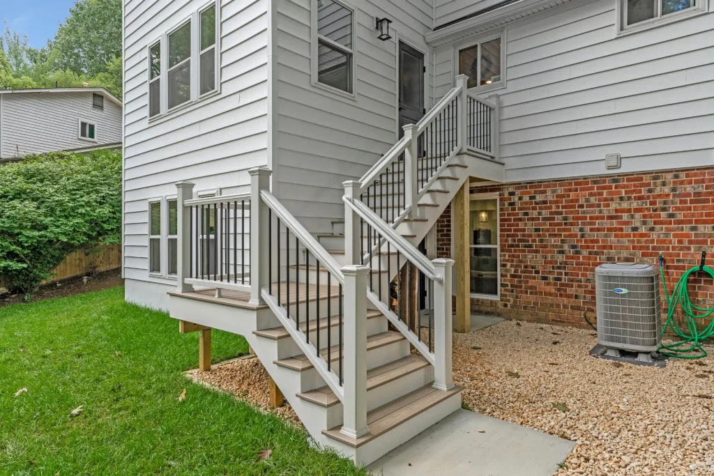 Two sets of outdoor stairs with white railings lead to a house with gray siding and red brick; a grassy yard and an air conditioning unit are visible.