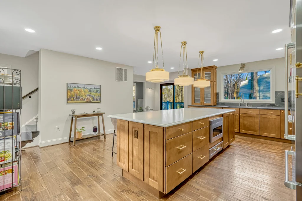 Modern kitchen with wooden cabinets, large island, pendant lights, stainless steel appliances, and hardwood floors. A window and sliding glass doors provide natural light.