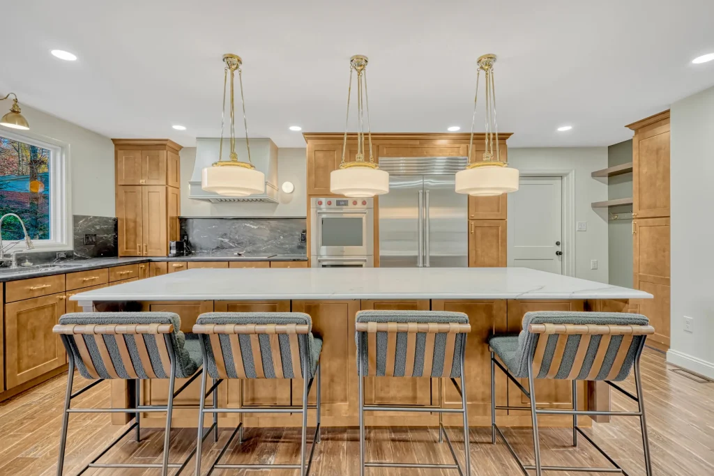Modern kitchen with wooden cabinets, a large island with white countertop, four bar stools, pendant lights, and stainless steel appliances.