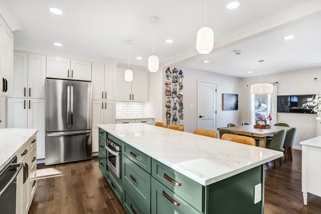 Modern kitchen with a large marble island, green lower cabinets, stainless steel appliances, white upper cabinets, and an adjacent dining area with a wooden table and yellow chairs.