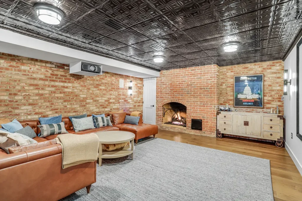 Basement living room with exposed brick walls, a black patterned ceiling, fireplace, sectional sofa, round ottoman, gray rug, and a vintage-style sideboard with framed poster.