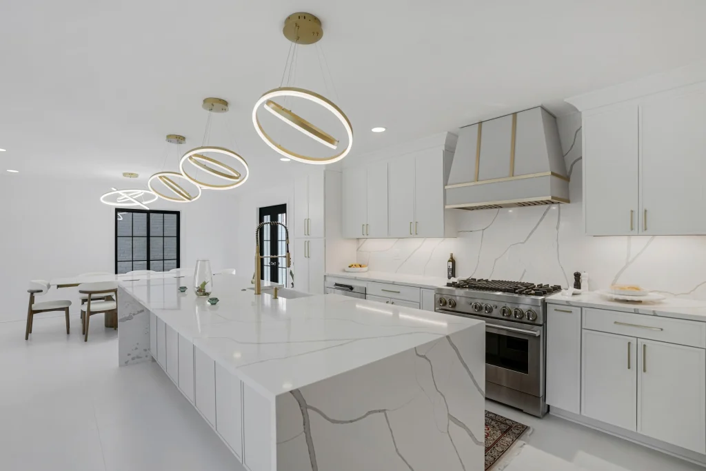 Modern kitchen with white cabinets, marble countertops and backsplash, stainless steel appliances, and circular pendant lights above a large island. Dining area visible in the background.