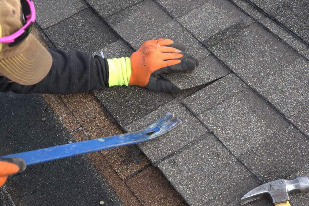 man using pry bar to remove shingles; essential roofing tools